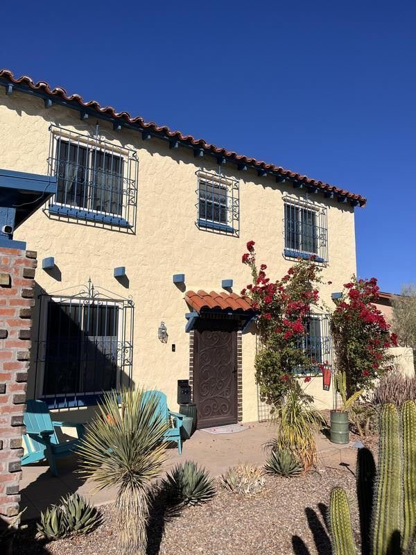 A Spanish-style house with stucco walls, terracotta roof tiles, desert landscaping, vibrant bougainvillea, turquoise chairs, and wrought iron accents in Tucson's arid environment.