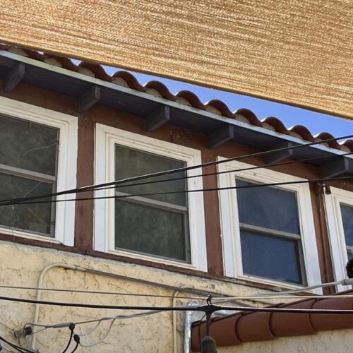 A Tucson building with white-framed windows under a woven awning supported by rustic wooden beams, terracotta roof tiles, utility wires, and a hanging bulb showcasing historic and urban influences.