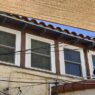 A Tucson building with white-framed windows under a woven awning supported by rustic wooden beams, terracotta roof tiles, utility wires, and a hanging bulb showcasing historic and urban influences.