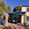 Charming Tucson residential scene featuring a carport with brick pillars and a blue roof, a sandy yellow Spanish-style house, bins on a brick driveway, lush tree, and drought-tolerant plants.