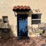 A serene Southwest patio features a warm stucco wall with a vibrant blue door and terracotta awning, surrounded by rustic bricks and shaded greenery, evoking a peaceful Tucson charm.