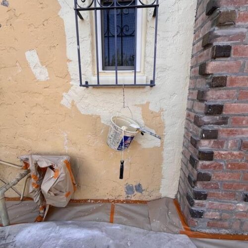 A textured wall undergoing the Terrenella Painting Project shows contrasting painted and unpainted areas, a paint can with a brush, protective plastic sheeting, and a black metal grille window.