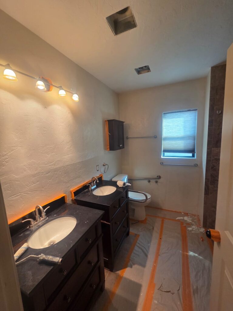 Interior painting in Tucson bathroom with fresh neutral walls, dark vanities, black countertops, and protective floor sheeting during renovation work.